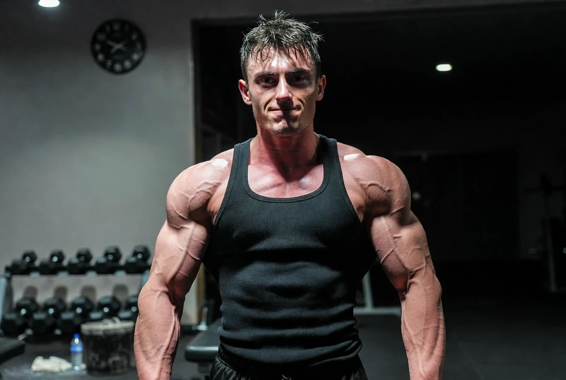 Schlanker, muskulöser Bodybuilder in einem Fitnessstudio, der Anzeichen von Nebenwirkungen von Trenbolonacetat aufweist: dunkle Augenringe aufgrund von Schlaflosigkeit, leichter Schweiß aufgrund von nächtlichen Schweißausbrüchen und erhöhtem Stoffwechsel sowie angespannter Gesichtsausdruck – trotz extrem harter Muskeln und geringem Körperfettanteil.