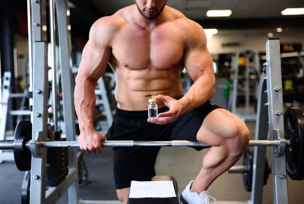 Sportlicher Mann in einem Fitnessstudio, der eine Ampulle Testosteron-Enanthate in der Hand hält, mit einem Notizblock, auf dem die wichtigsten Wirkungen (Muskelaufbau, Energie, Libido) und Nebenwirkungen (erhöhter Östrogenspiegel, niedrigerer HDL-Spiegel) sowie ein Bluttestbericht zu sehen sind – zur Veranschaulichung der tatsächlichen physiologischen Veränderungen nach der Injektion.